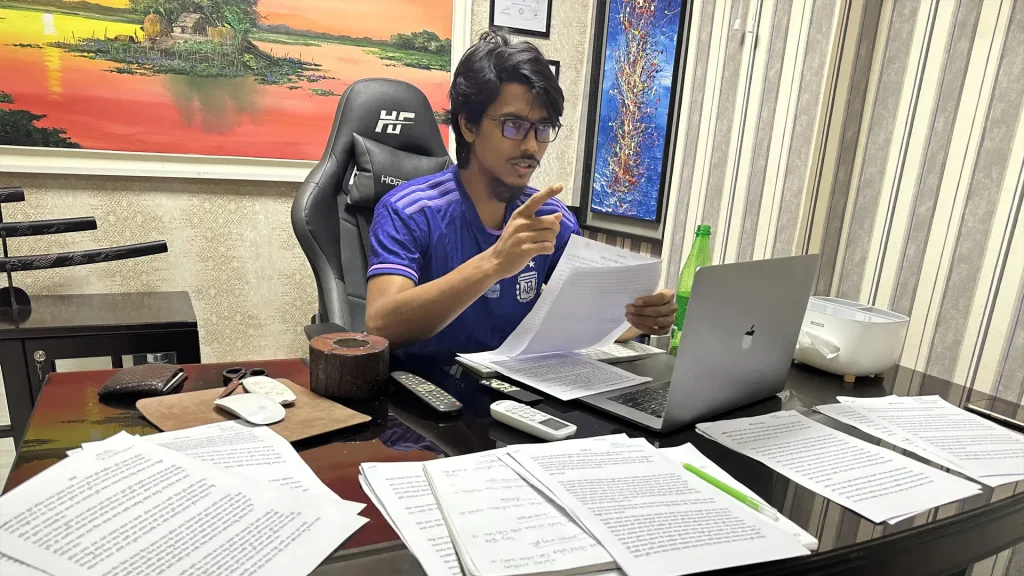 Faisal Khan Shuvo reviewing printed scripts at a desk with a laptop